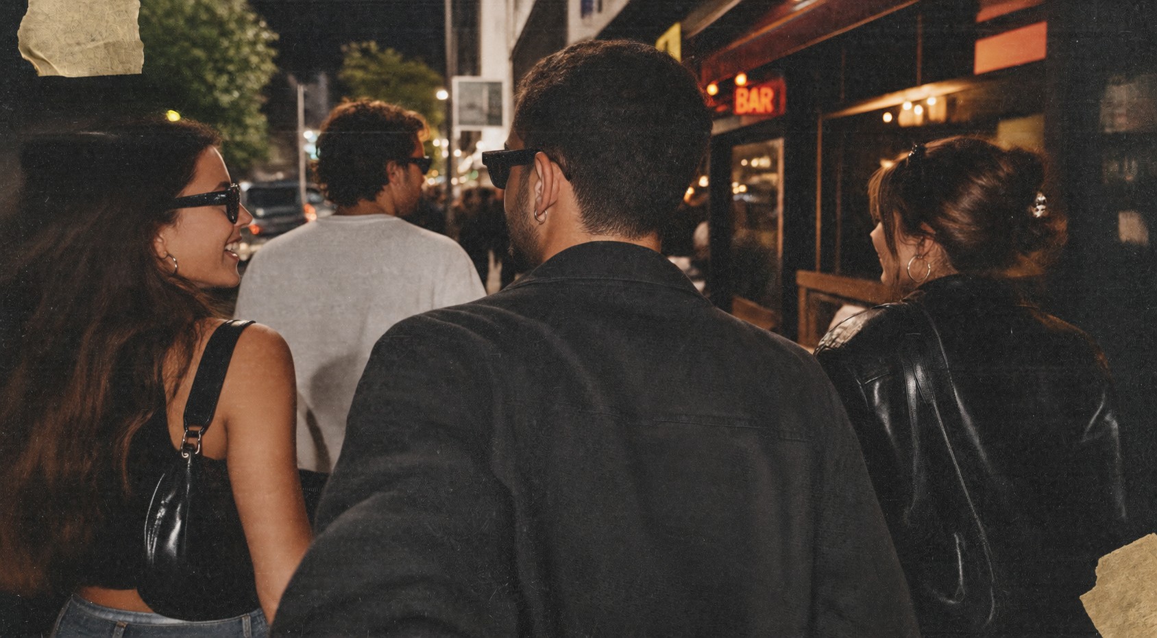 Four friends walking down a city street at night, past a bar