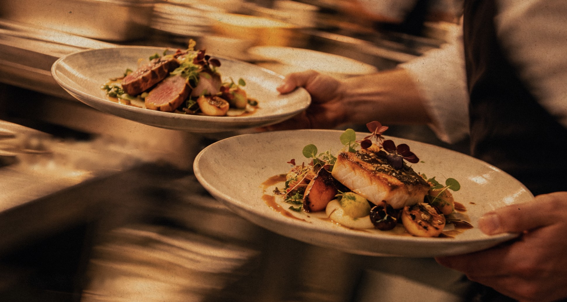A chef carrying two plated dishes through a busy kitchen